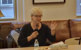 Jenny Van Hook, a white woman with blonde hair and glasses in a dark shirt, talks into a microphone.