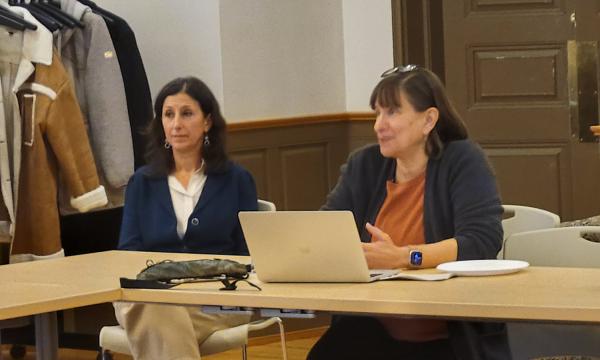 Deborah Ehrenthal, a white woman in a dark sweater and orange shirt with short hair, addresses the group while sitting next to Jennifer Glick, a white woman with a dark colored sweater, white shirt and light-colored pants.