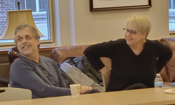 Marty Sliwinski, a white man with grey hair in a blue sweater, smiles while sitting next to Jenny Van Hook, a white woman with blonde hair and glasses in a black sweatter.