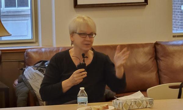 Jenny Van Hook, a white woman with blonde hair and glasses in a dark shirt, talks into a microphone.