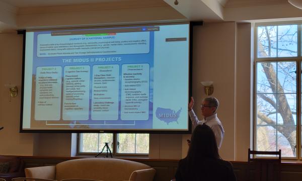 Dave Almeida, a white man in white shirt and brown pants, presents about working with MIDUS data.