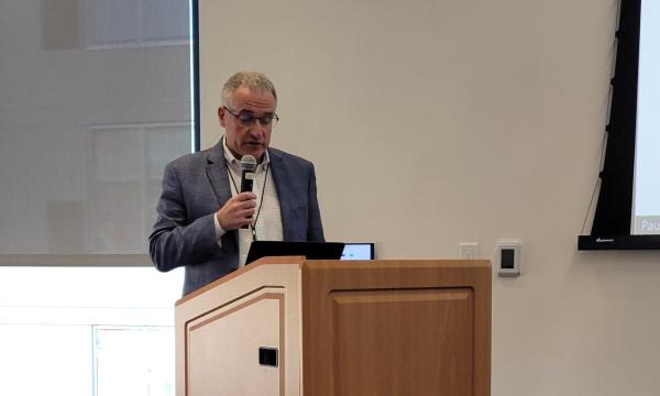 Andrew Read, an older man in a blue jacket, dark pants, and white collared shirt, holds a microphone and stands at a light wood colored podium.