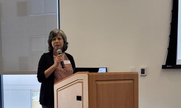 Rina D. Eiden, a middle aged woman with short hair, wearing a black sweater and red patterned shirt, stands at the podium and holds a microphone.
