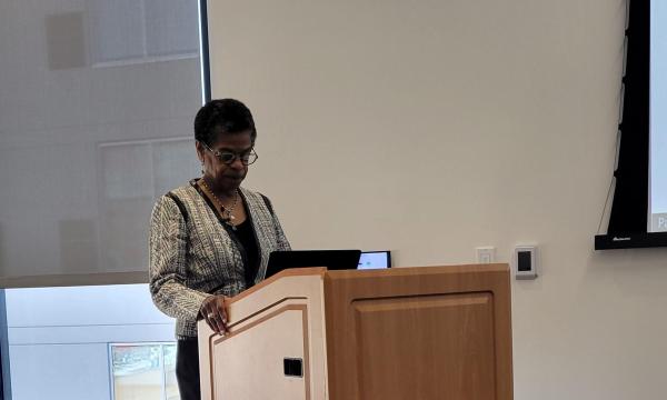 R. Lorraine Collins, a middle-aged black woman in a white and black patterned jacket and black pants, stand at the light wood colored podium.