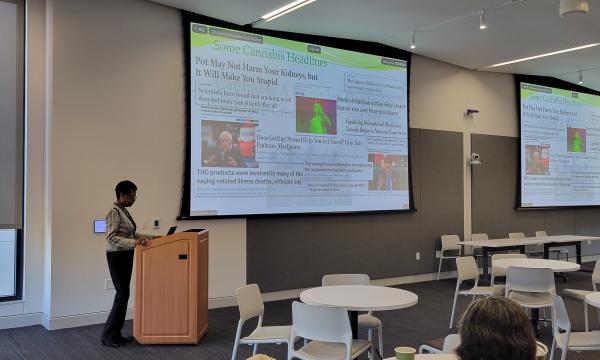 R. Lorraine Collins, a black woman wearing a black and white jacket with black pants, stands at a light wood colored podium next a screen with her keynote PowerPoint.