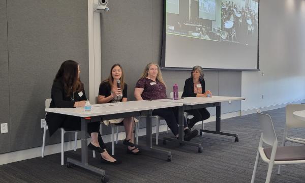 Four middle-aged white women sit at a table in front of CSUA conference attendees.