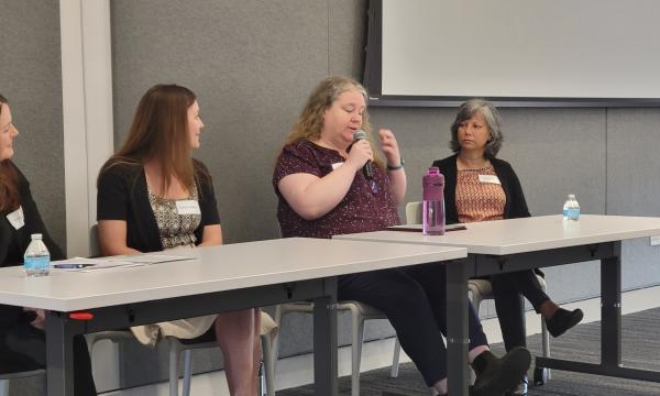 Four middle-aged white women sit at a table in front of CSUA conference attendees.