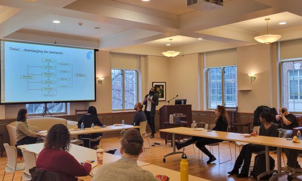 Mohanraj Krishnan, a faculty member wearing a long cardigan and white button-up, presents to the group in a wide shot.
