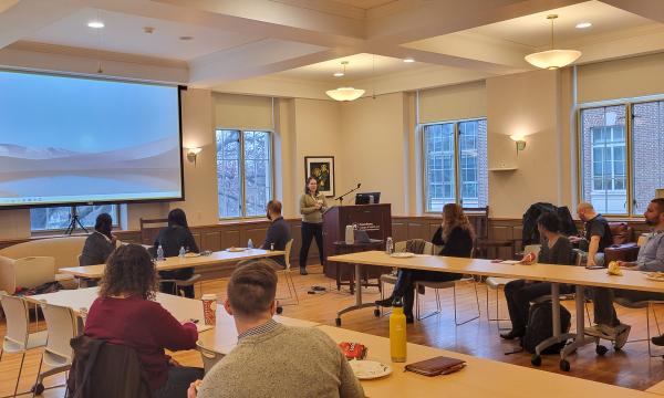 Tracee Saunders, a woman with shoulder-length brown hair wearing a green sweater, presents to the group in a wide shot. 
