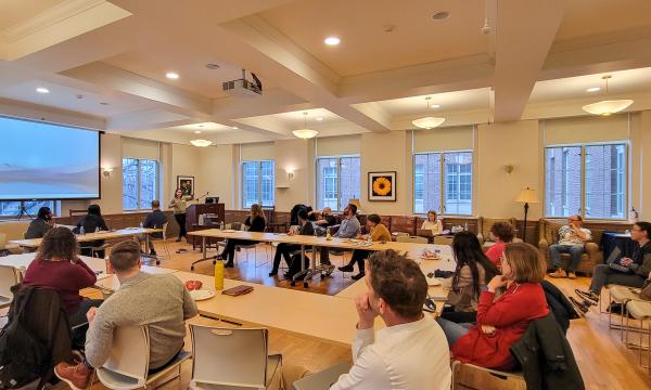Tracee Saunders, a woman with shoulder-length brown hair wearing a green sweater, presents to the group in a wide shot. 