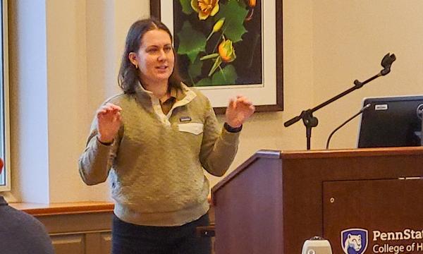 Tracee Saunders, a woman with shoulder-length brown hair wearing a green sweater, presents to the group. 