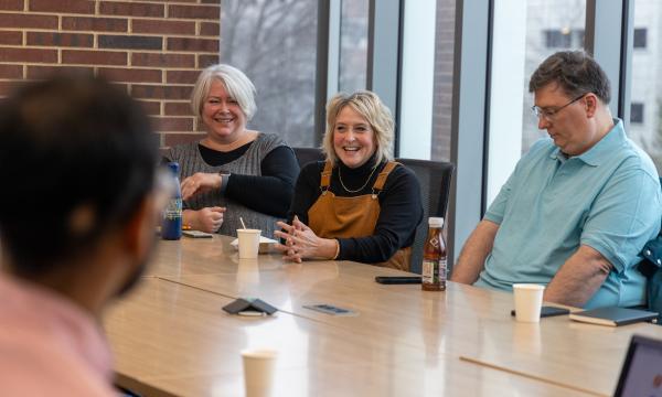 Three people sit at a conference table, a woman in a grey vest with white hair, a woman with blonde hair wearing brown overalls, and a man in a blue colored polo shirt.