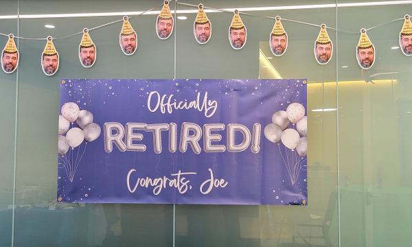 Blue sign that says "Officially Retired! Congrats Joe" on it with a banner of Joe's face hanging above it.